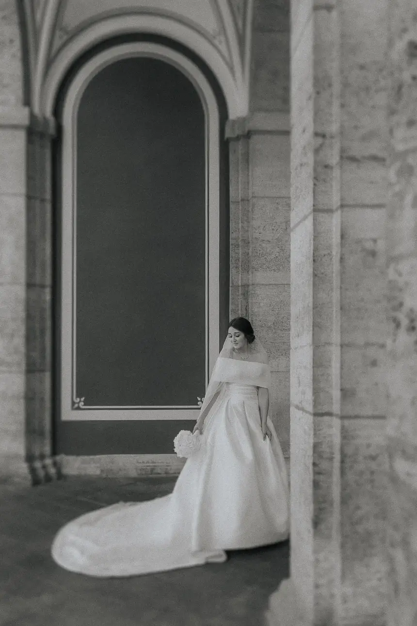 schwarz weiss Foto einer edlen Braut in München die sich in einem Säulengang bewegt und auf Ihren Brautstrauß blickt